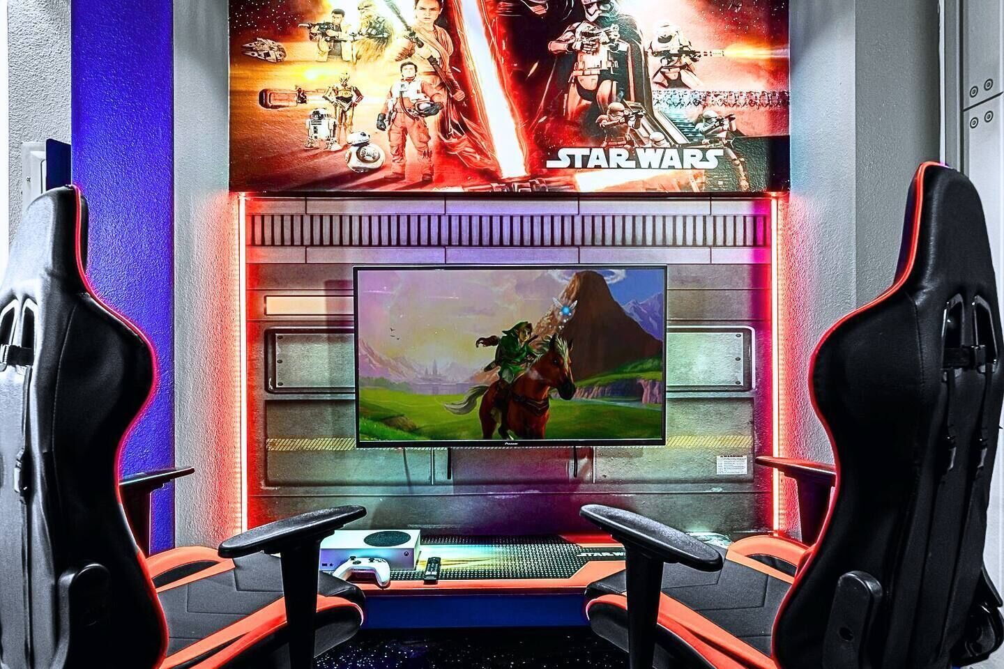 Un espacio para videojuegos con dos sillas ergonómicas negras, una pantalla grande mostrando una película y una pared temática de Star Wars.