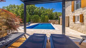 Un espace piscine extérieur moderne, doté de deux chaises longues, d’un bâtiment en pierre et d’un ciel d’un bleu limpide.
