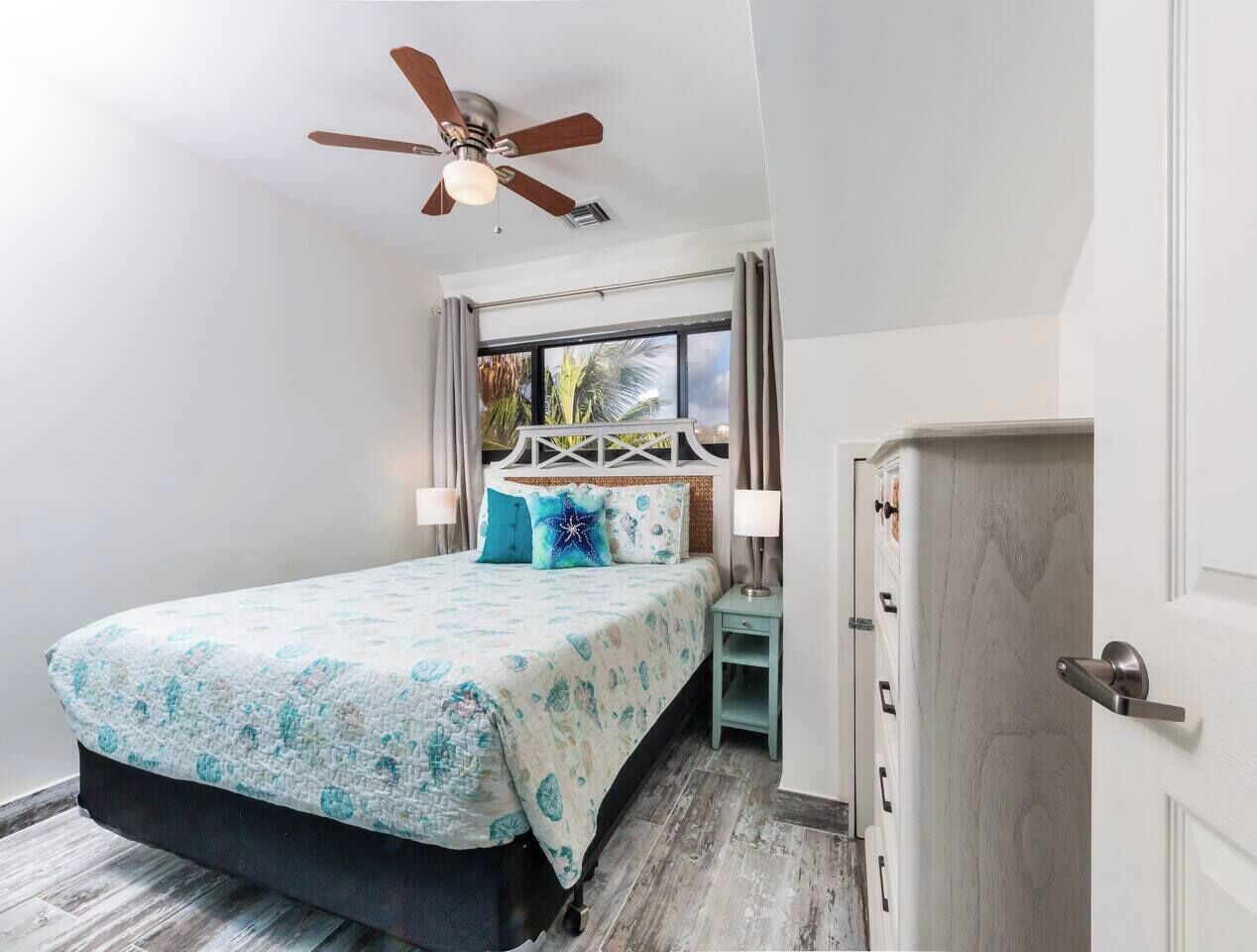 Un dormitorio con cama, ventilador de techo, mesita de noche, cómoda y ventana con cortinas.