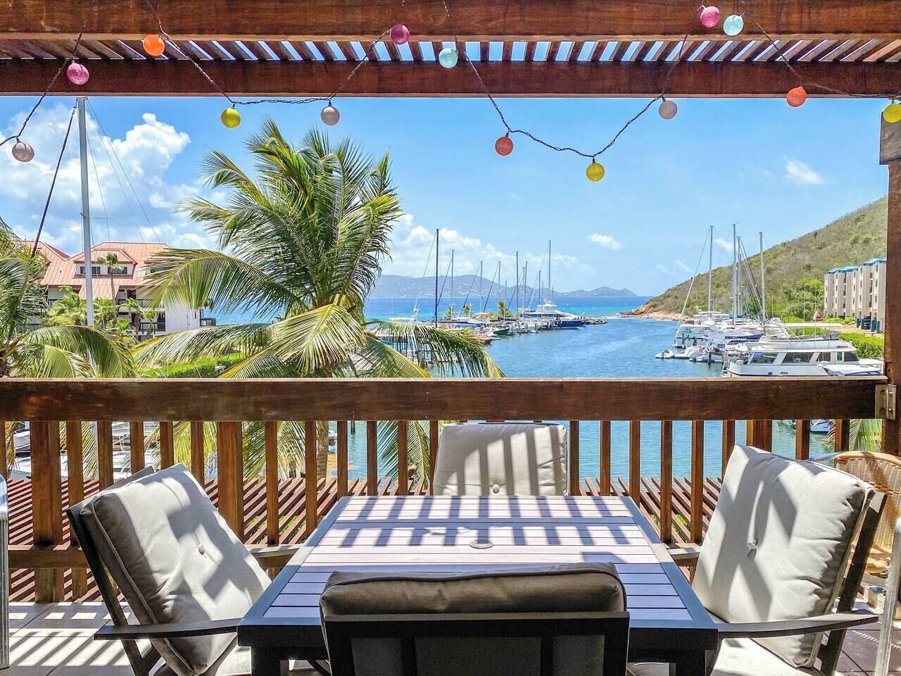 A wooden deck with a table and chairs, overlooking a marina with boats and a clear blue sky.