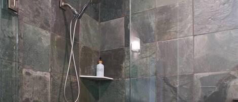 A shower with a glass door, a handheld showerhead, and a shelf with a bottle.