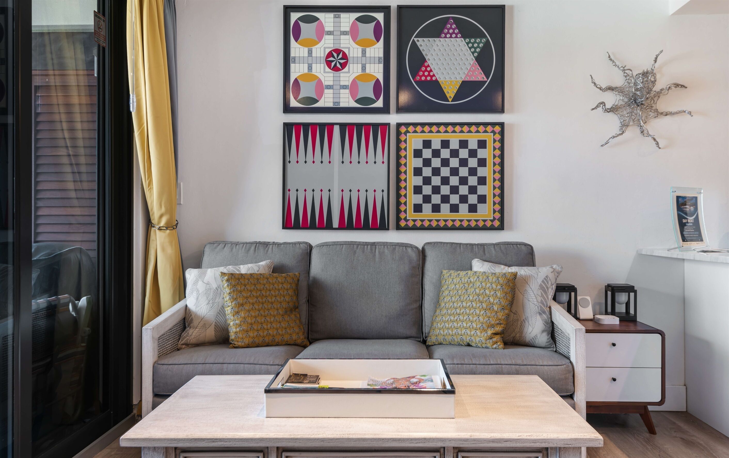 A living room with a grey sofa, patterned cushions, a coffee table with a magazine, and four framed artworks on the wall.