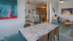 Un coin repas avec une table en marbre dressée pour six personnes, entourée de chaises de couleur turquoise. On aperçoit au fond une cuisine avec des meubles en bois et un ventilateur de plafond.