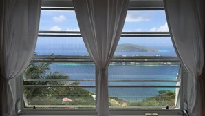 Une chambre dotée d’une grande fenêtre donnant sur la côte, avec un lit et des rideaux.