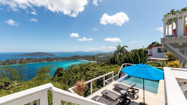 Un espace piscine doté de chaises longues et d’un parasol, offrant une vue sur une mer turquoise et un paysage montagneux.