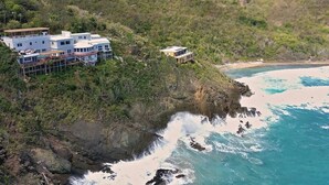 Una propiedad costera con varias edificaciones, incluyendo una casa grande y una estructura más pequeña, ubicada en un acantilado con vistas a una playa y el océano.