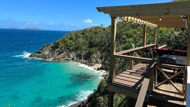 Una terraza de madera con vistas al mar turquesa, una costa rocosa y abundante vegetación.