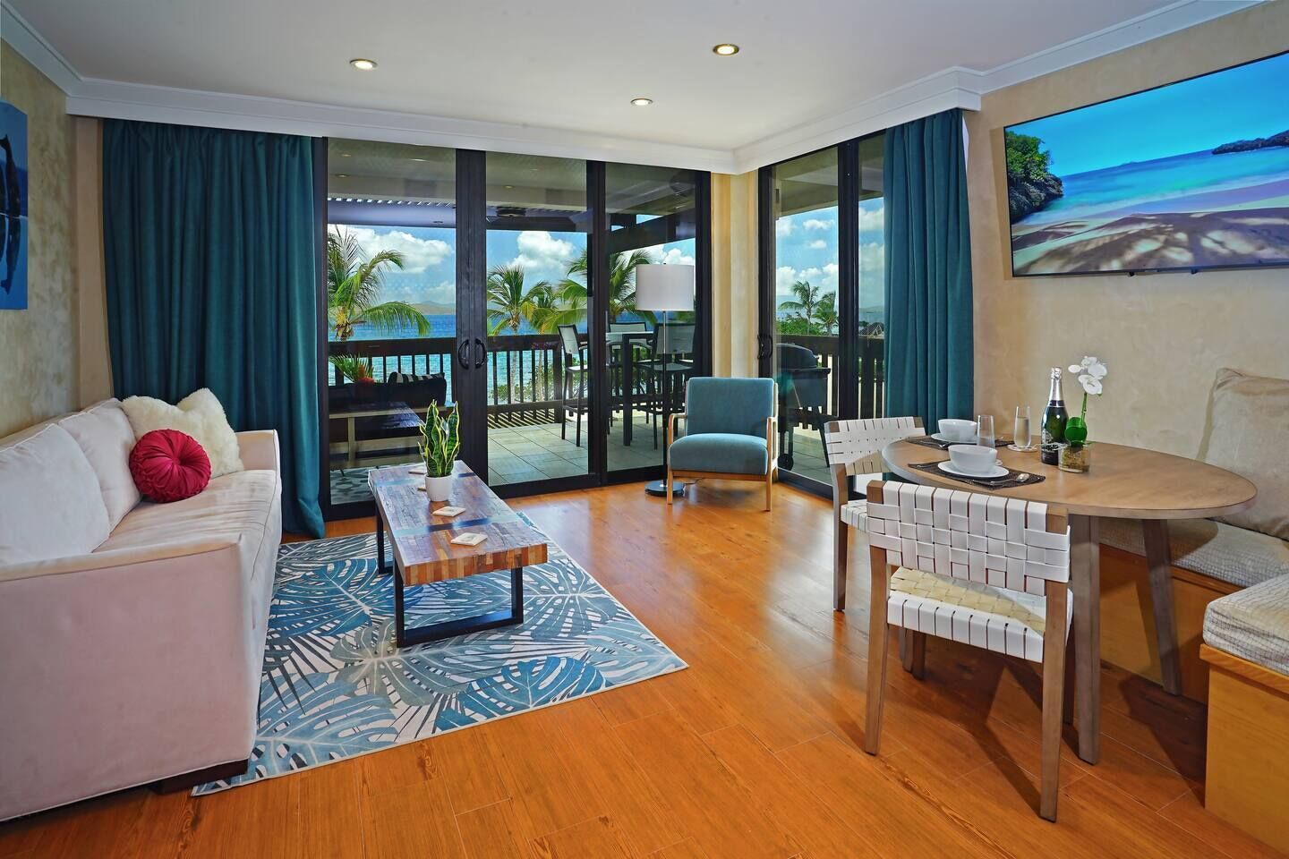 A modern hotel room with a balcony, a sofa, a coffee table, a dining table, and a painting of a beach scene.