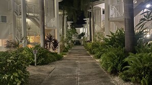 Vue nocturne d’un couloir d’hôtel, avec une allée centrale bordée de palmiers et de verdure, menant à un bâtiment doté de nombreux balcons et d’escaliers.