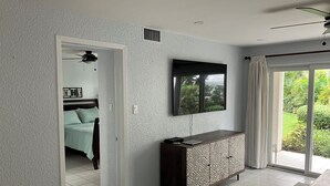 A modern living room with a flat-screen TV, a dark-colored sofa, and a patterned cabinet. A door leads to a bedroom with a bed and a ceiling fan. - Resort style ground floor  condo steps from the beach  (Georgetown)