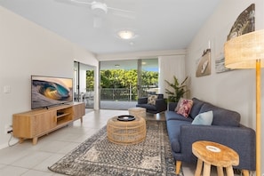 A modern living room with a flat-screen TV, a blue sofa, a wicker coffee table, and a wooden stool. - Beach Bliss - 3 Bedroom Apartment (Kingscliff)