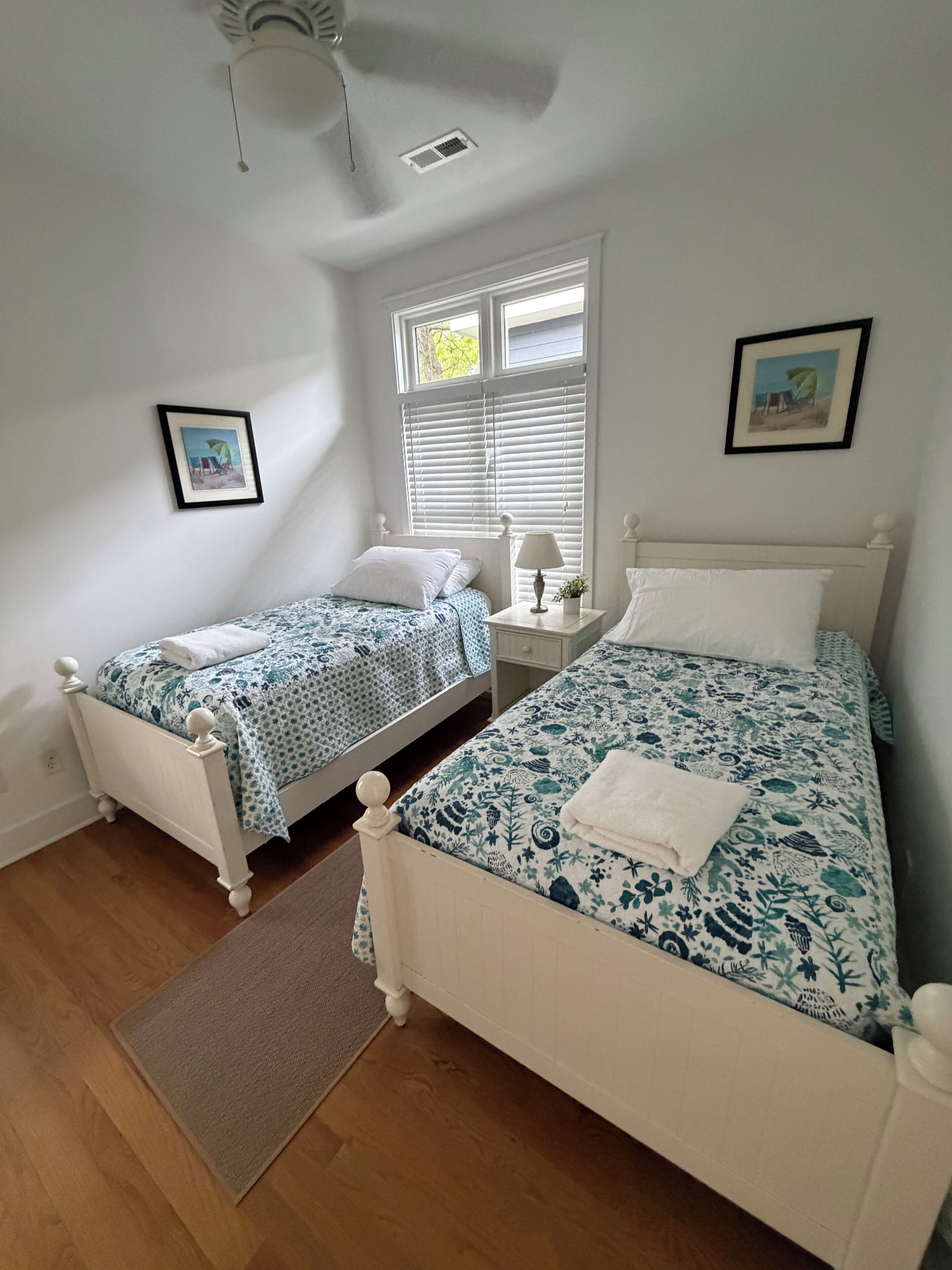 A bedroom with two beds, a window with blinds, a ceiling fan, and a small bedside table with a lamp.