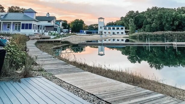 Un muelle de madera que conduce a un lago tranquilo, rodeado de casas y vegetación.