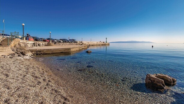 Una platja de petits còdols amb un moll, aigua blau clar i muntanyes allunyades.
