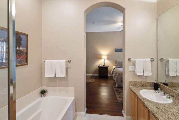 Un baño de hotel con una bañera, un lavabo y un espejo. Hay un pasillo que conduce a un dormitorio con una cama y una mesita de noche con una lámpara.