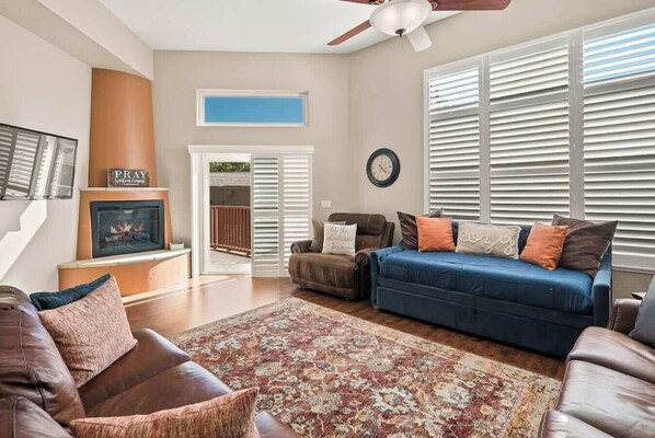Un amplio salón con chimenea, ventilador de techo y un gran ventanal con persianas.