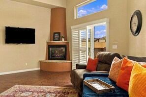 Una sala de estar con chimenea, un televisor de pantalla plana montado en la pared, un sofá con cojines de colores y una ventana con persianas.