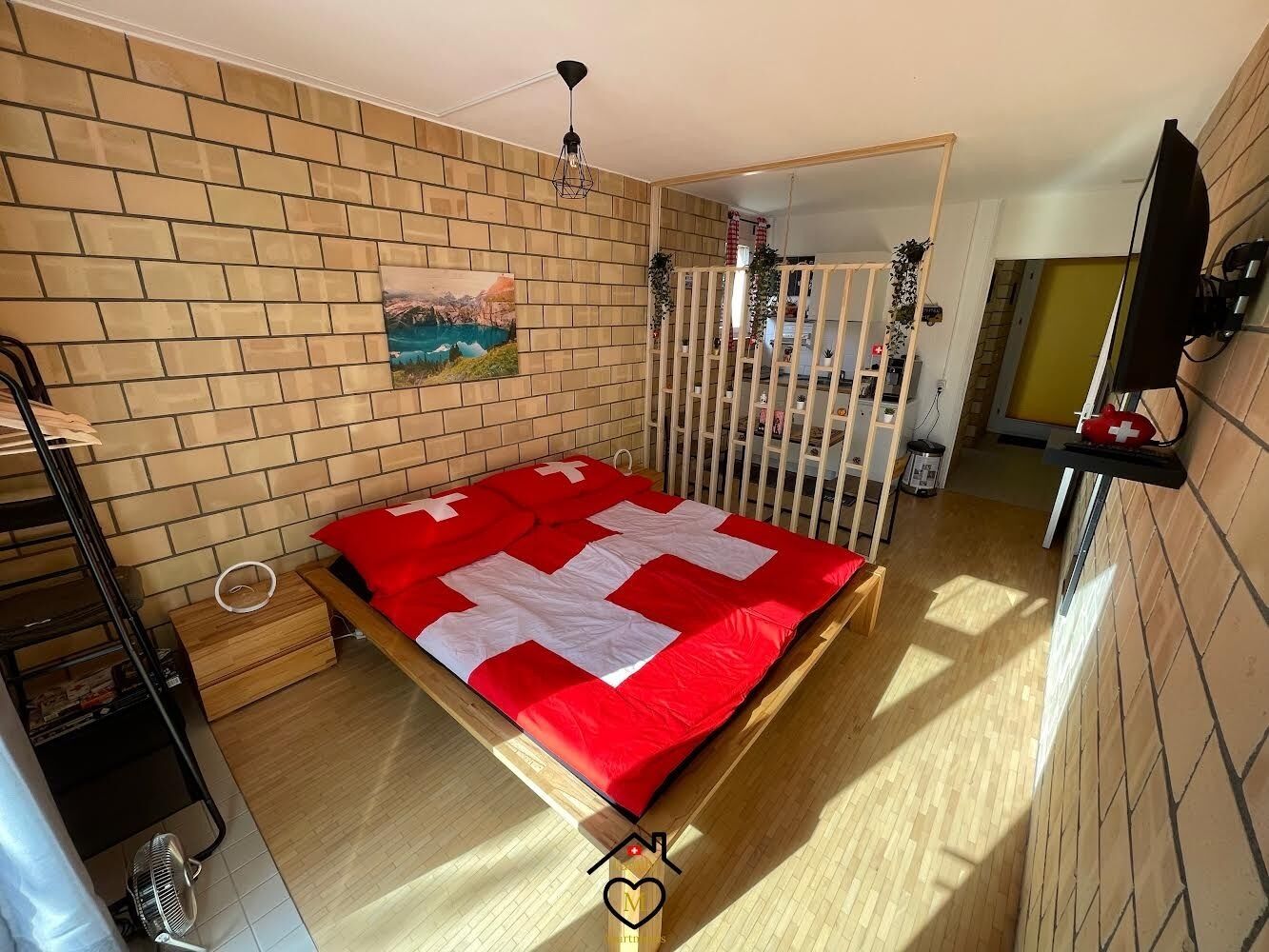 A compact bedroom with a bed, a wooden floor, a brick wall, a painting, a white crib, and a TV on a shelf.