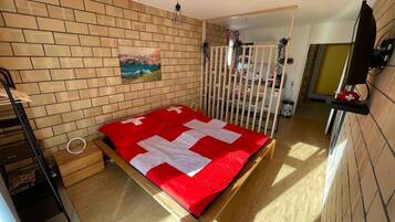 A compact bedroom with a bed, a wooden floor, a brick wall, a painting, a white crib, and a TV on a shelf.