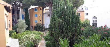 Une cour intérieure avec un chemin pavé, entourée de bâtiments dotés de balcons et agrémentée de diverses plantations, notamment des arbres et des arbustes.