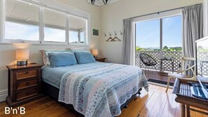 A bedroom with a bed, bedside tables, a chandelier, a balcony with chairs, and a view of the outdoors. - Warm and welcoming, private one bedroom on a lifestyle property. (Waingawa)