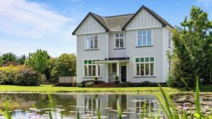 A two-story house with a front porch, a pond in the foreground, and a garden with various plants and flowers. - Warm and welcoming, private one bedroom on a lifestyle property. (Waingawa)