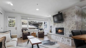A modern living room with a fireplace, a television mounted on a stone wall, and a view of a balcony with outdoor furniture.