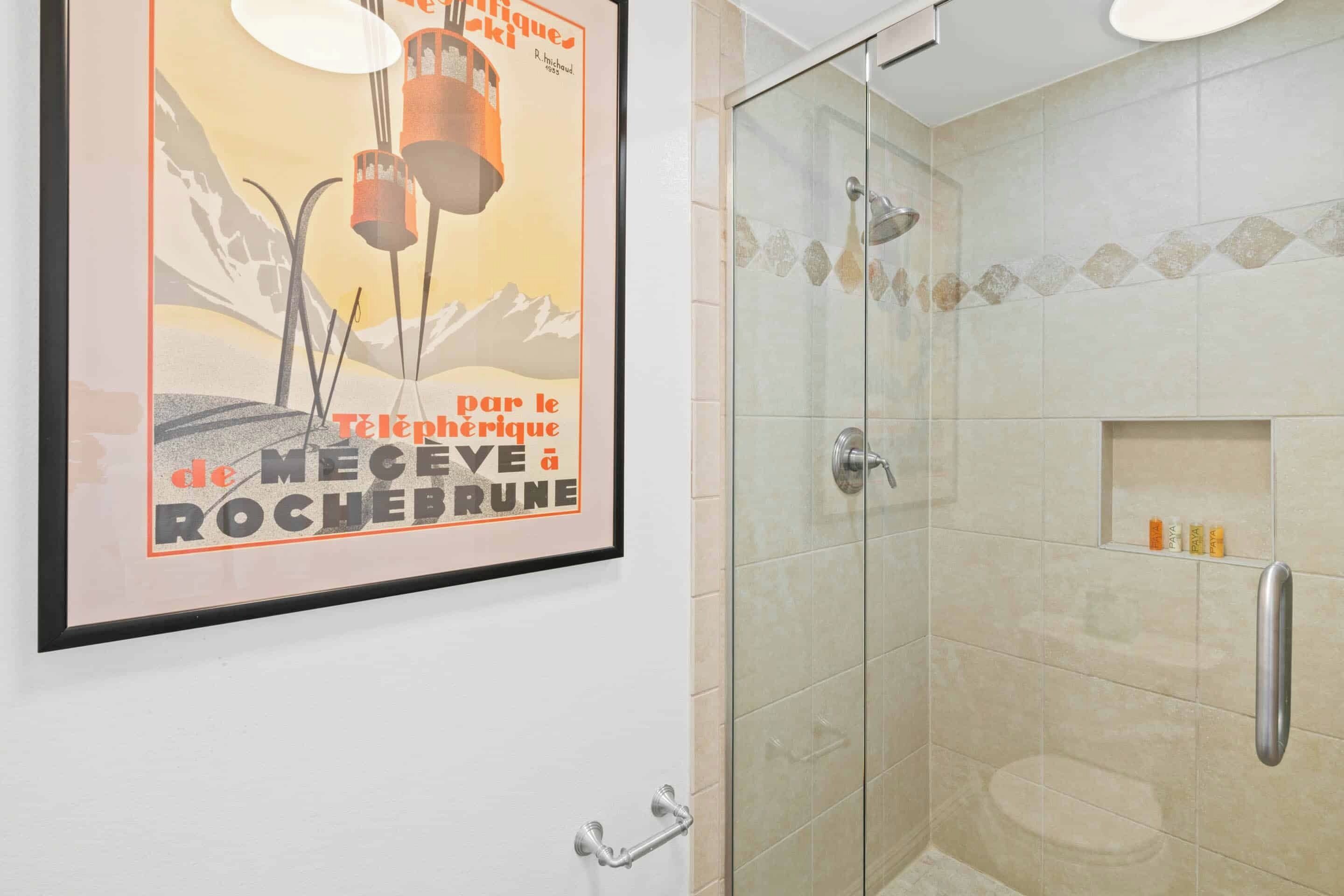 A framed poster with a mountain and hot air balloon, a glass-enclosed shower area with a towel rack, and a shelf with toiletries.