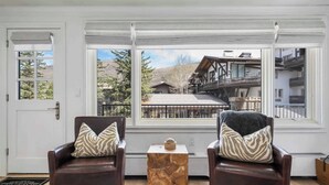 A room with two leather armchairs, a wooden coffee table, and large windows offering a view of a chalet.