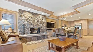 A spacious living room with a stone fireplace, a dining area with a wooden table, and a kitchen with wooden cabinets and a chandelier.