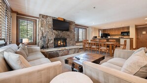 A spacious living area with a stone fireplace, comfortable seating, a dining table, and a kitchen in the background.