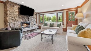 A living room with a stone fireplace, a flat-screen TV, a sofa, a coffee table, and large windows with a view of greenery. - Lodge Tower 294 2BR at the base of Vail Mountain (Vail)