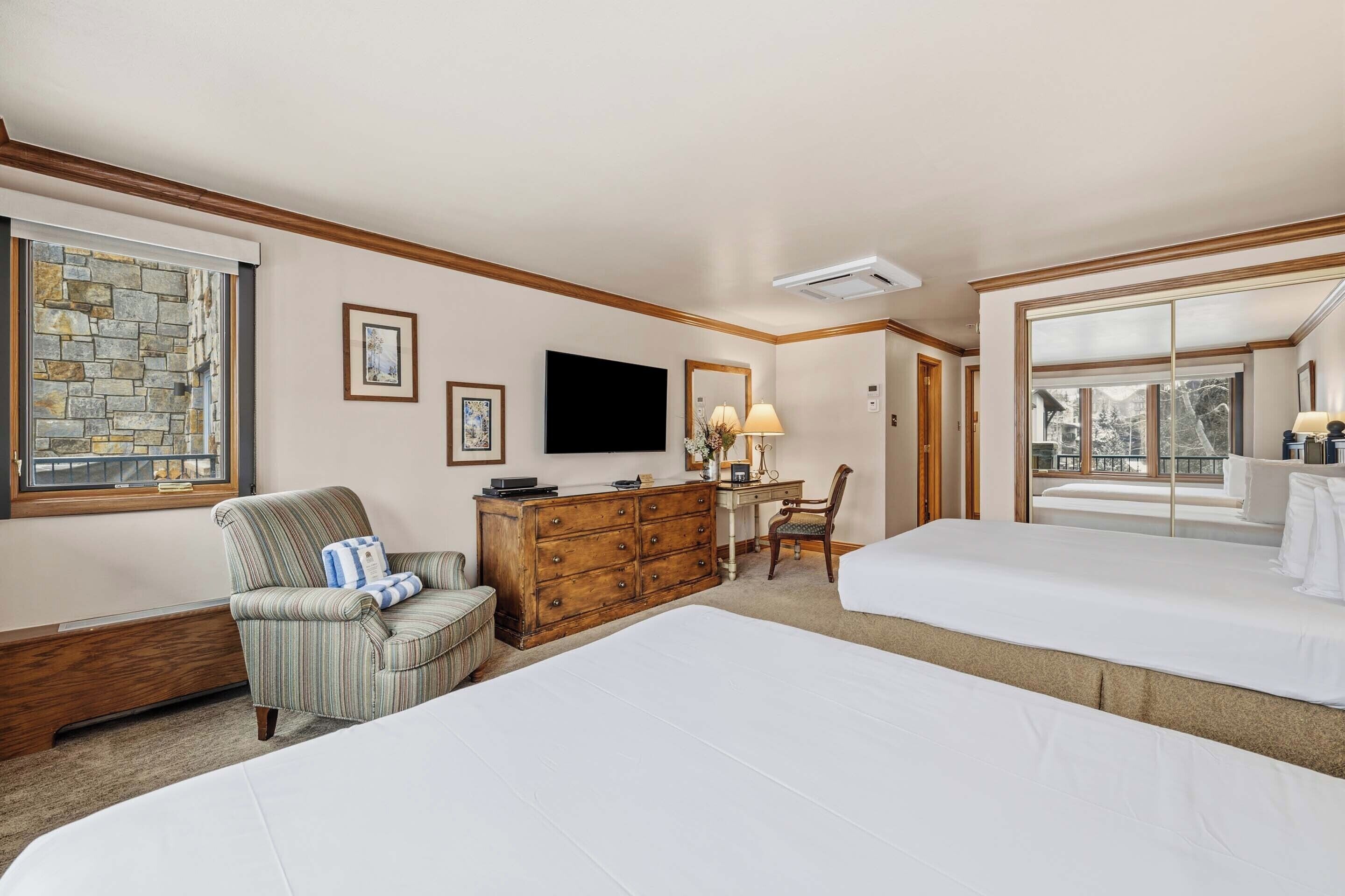 Een hotelkamer met twee bedden, een houten dressoir, een bureau, een stoel, een televisie en een groot raam.