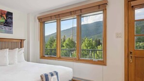 Une chambre avec un grand lit, un banc et une fenêtre donnant sur des arbres et des montagnes.