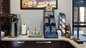 Una kitchenette de hotel con una cafetera, dispensadores My Coffee y un cartel de My Place que anuncia desayuno en la cama.