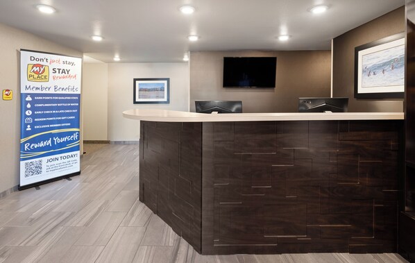 A hotel reception area with a dark wood reception desk, a framed picture on the wall, and a signboard advertising member benefits.