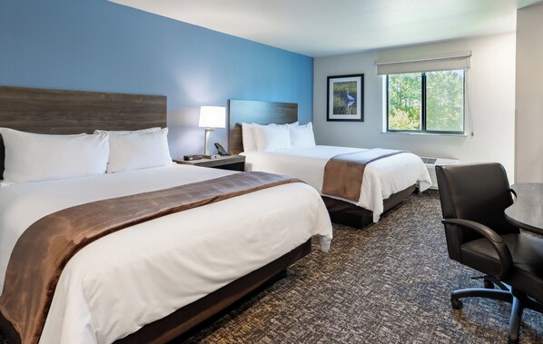 A hotel room with two beds, a desk, a chair, and a window with a view of trees.