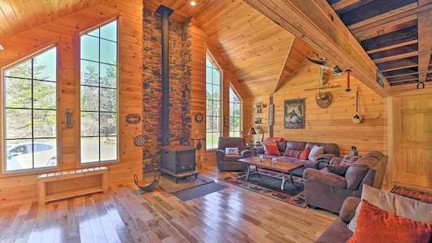 A log cabin interior with a stone fireplace, wooden walls, large windows, and a cozy seating area. - Unadilla Cabin on 50 Acres + Fire Pit & Creek (Unadilla)