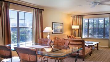 A hotel room with a glass table, two patterned armchairs, a sofa, a lamp, and a framed picture on the wall.
