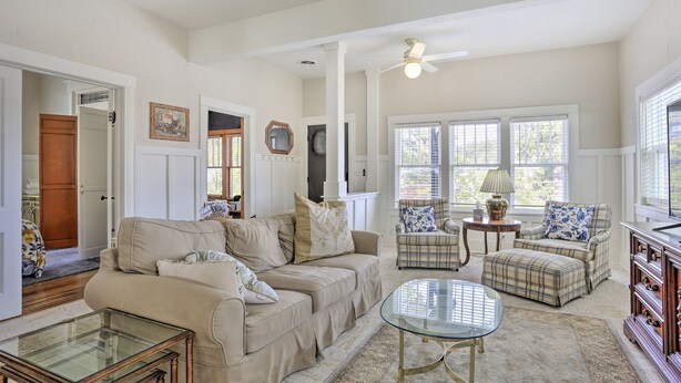 A spacious living room with a beige sofa, a plaid armchair, and a glass coffee table. There is a wooden cabinet with wine bottles and a ceiling fan. - Mountain-View Lead Home w/ Patio + Fire Pit! (Lead)