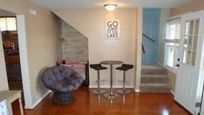 A living room with a grey bean bag chair, two bar stools, a small table, and a staircase leading to a door. - Charming Lake View Cabin Retreat Near Captain Ron's Resort, Lake of the Ozarks (Sunrise Beach)