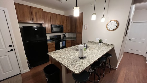 Una cocina con armarios de madera, un refrigerador negro, un microondas, una estufa y un reloj en la pared. Hay tres taburetes de bar y un cubo de basura.