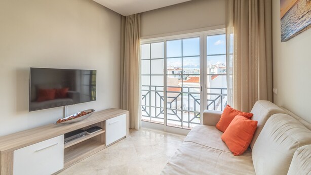 Ein modernes Wohnzimmer mit Flachbildfernseher, einer Couch mit orangefarbenen Kissen, einem Holztische und einem großen Fenster mit Blick auf Gebäude.