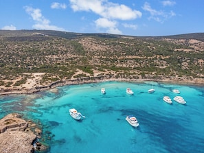 Una zona costera con barcos anclados en aguas azul turquesa, rodeada de terreno rocoso y vegetación escasa.