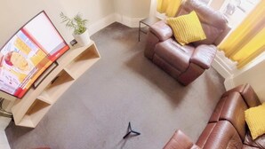 Zona salotto circolare con divani in pelle marrone e cuscini gialli, un tavolino con una pianta e un televisore su un mobile.