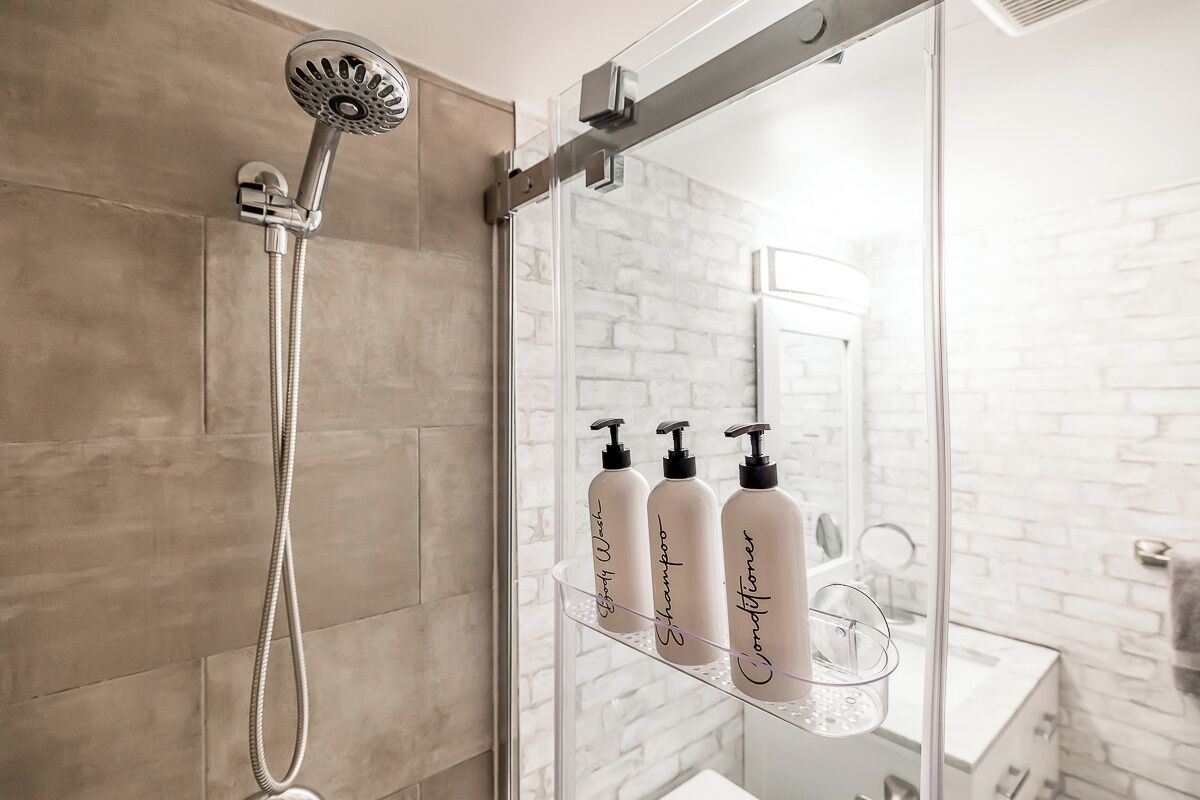 A shower with a handheld showerhead, a glass shower enclosure, and a glass shelf with three bottles of shampoo and conditioner.