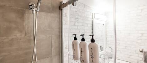 A shower with a handheld showerhead, a glass shower enclosure, and a glass shelf with three bottles of shampoo and conditioner.