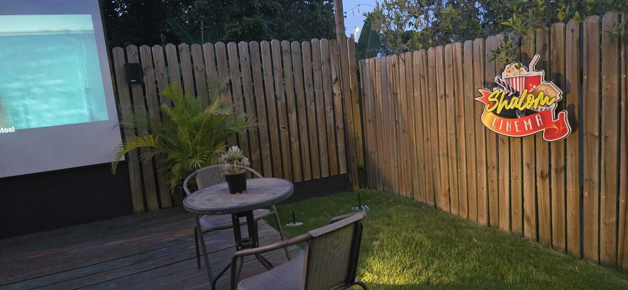Un patio trasero con una terraza de madera, una mesa redonda con sillas, una planta en maceta y una pantalla de proyección.