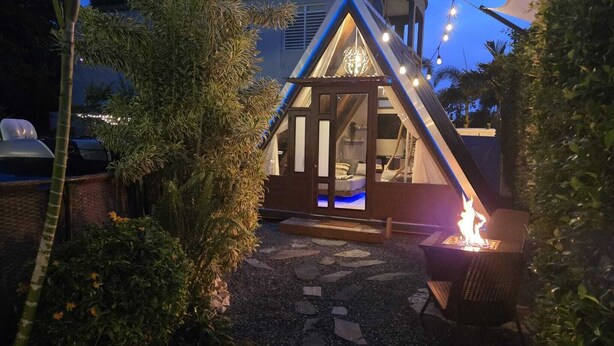 A cozy outdoor seating area with a fire pit, surrounded by lush greenery and a small building with a glass door. - A unique way to camp in luxury (Aguadilla Pueblo)