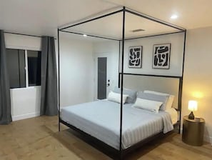 A four-poster bed with a white mattress and grey bedding, a bedside lamp, and abstract wall art.